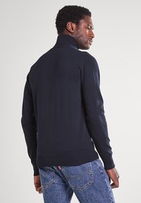 Granatowy sweter z wysokim kołnierzem, fakturą prążkowaną i długimi rękawami. Noszony na niebieskich dżinsach, podkreślający dopasowaną sylwetkę.