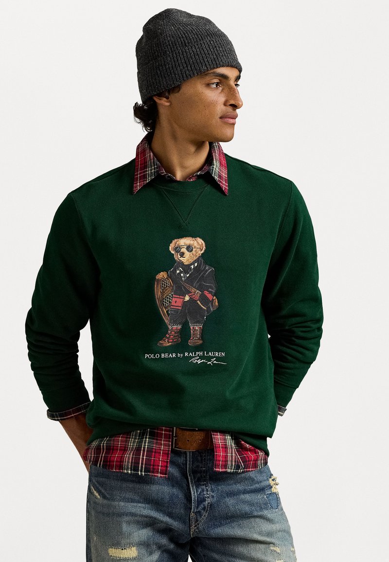 Zaļš džemperis ar lāča grafiku uzvalkā, uzvilkts virs sarkanā rūtainā krekliņa. Pelēka adīta beanie cepure, nolietoti zili džinsi, brūna josta.