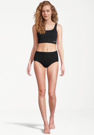 Junge Frau mit lockigem Haar, die ein schwarzes Crop-Top und schwarze High-Waist-Unterwäsche trägt, steht barfuß vor weißem Hintergrund.