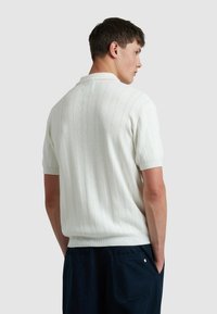 Kurzärmliges, weißes Strick-Polohemd mit vertikalen Rippungen, klassischem Kragen und strukturiertem Muster. Getragen mit einer navyblauen Hose, von hinten betrachtet.