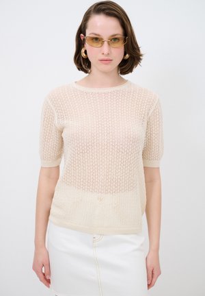Beige gebreide trui met een lossere weving, korte mouwen en een geribbelde halslijn. Gecombineerd met een witte denimrok voor contrast.