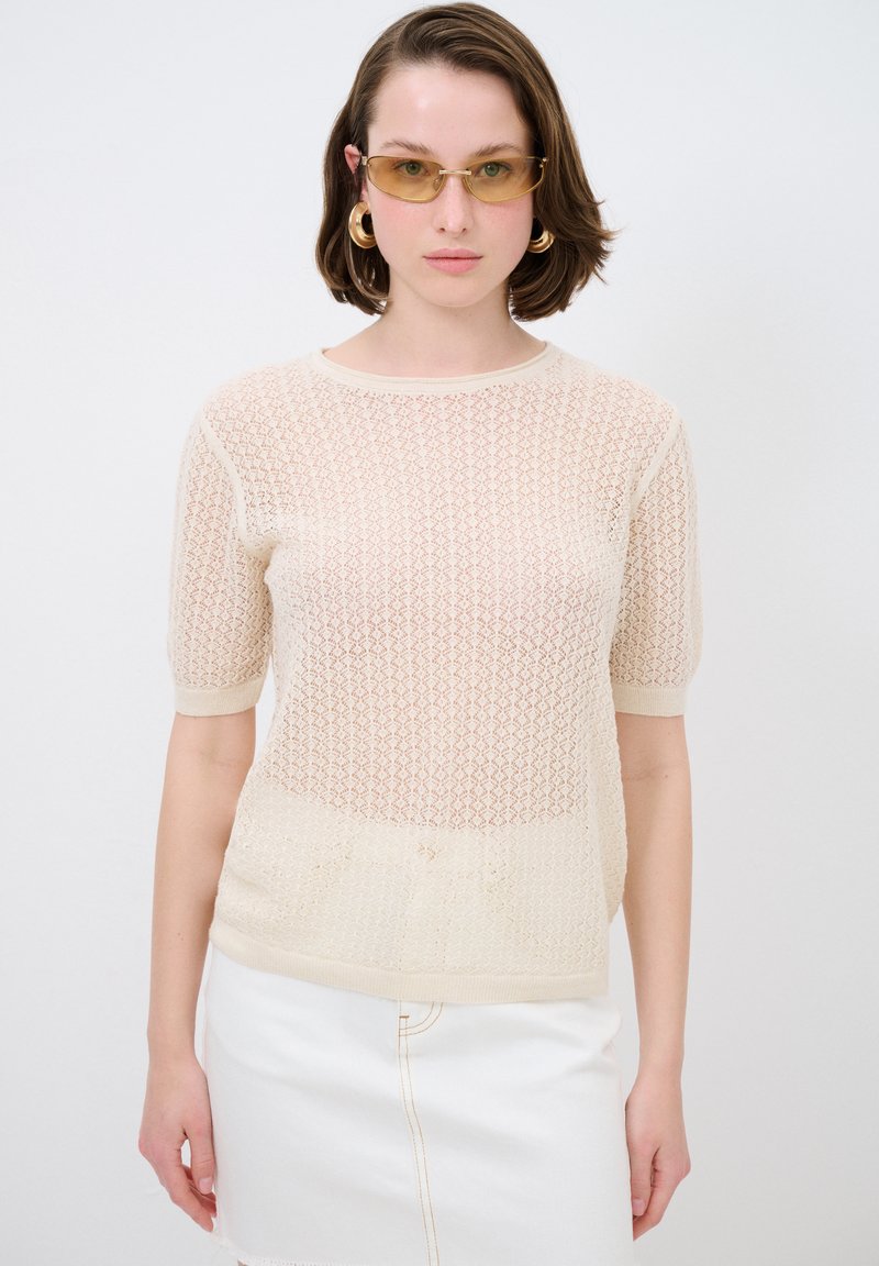 Beige Strickpullover mit lockerer Maschenstruktur, kurzen Ärmeln und geripptem Ausschnitt. Kombiniert mit einem weißen Denim-Rock für einen Kontrast.