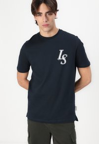 T-shirt en coton bleu marine arborant un logo LS blanc sur la poitrine gauche, à manches courtes et avec un col rond, dotée de fentes sur les côtés à l'ourlet.
