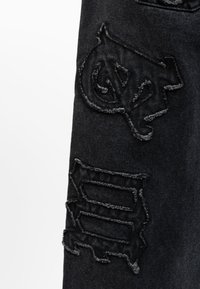 Jeans in denim nero con toppe strappate che presentano disegni ricamati. L'orlo evidenziato e i dettagli testurizzati arricchiscono l'interesse visivo del tessuto.