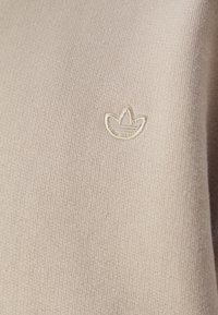 Ljust beige tyg med en liten, broderad logotyp i form av ett trebladigt blad i något mörkare beige tråd.