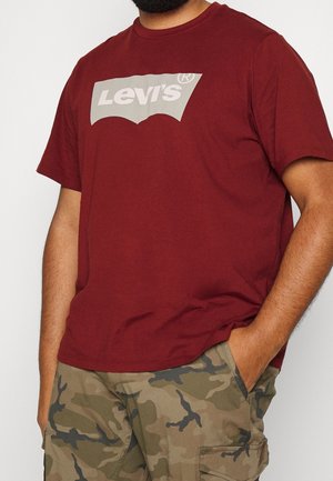 Mann trägt ein rotes Levi’s-T-Shirt und Tarn-Cargoshorts, steht mit einer Hand in der Tasche vor einem schlichten Hintergrund.