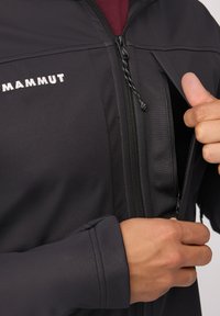 Zwarte softshell jas met een hoge kraag, ritszak en elastische manchetten. Beschikt over een opvallend wit logo op de borst voor branding.