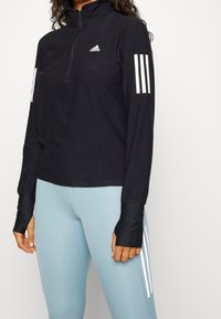 Femme portant un haut noir à manches longues Adidas avec des trous pour les pouces et des leggings bleu clair avec des bandes blanches sur le côté.
