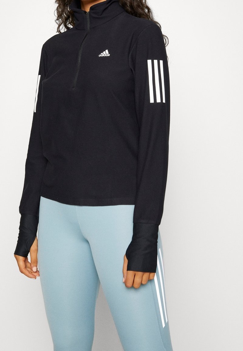 Femme portant un haut noir à manches longues Adidas avec des trous pour les pouces et des leggings bleu clair avec des bandes blanches sur le côté.
