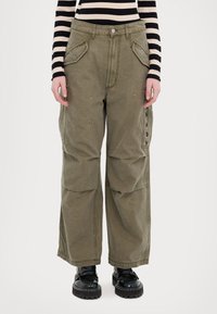 Pantaloni cargo khaki, dintr-un material moale, cu picioare largi, buzunare frontale mari și o cusătură decorativă pe exterior. Asortați cu pantofi platform negri.