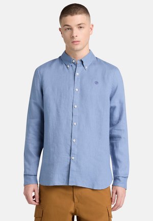 Giovane uomo che indossa una camicia azzurro chiaro con bottoni e un piccolo logo sul petto, pantaloni cargo marroni, in piedi davanti a uno sfondo semplice.