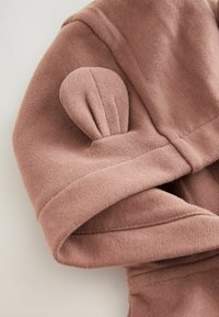 Tissu tricoté rose pâle, avec un détail en forme de volants arrondis sur la manche. Texture lisse avec un repli au poignet. Design subtil et minimaliste.