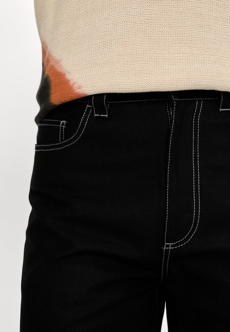 Schwarze Denim-Jeans mit kontrastierenden weißen Nähten, kombiniert mit einem beigen Strickoberteil mit orangefarbenen und schwarzen Batikdetails am Saum.