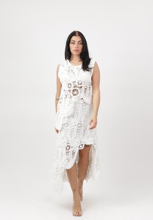 Frau mit dunklen Haaren, die ein ärmelloses weißes Spitzenoberteil und einen passenden High-Low-Rock trägt, barfuß vor einem einfarbigen Hintergrund posierend.