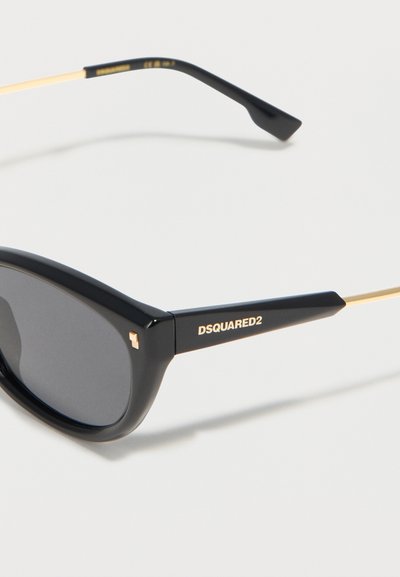Μαύρα γυαλιά ηλίου DSQUARED2 με οβάλ σκούρους φακούς, παχιά μαύρη μπροστινή πρόσοψη και λεπτούς χρυσούς μεταλλικούς βραχίονες σε λευκό φόντο.