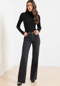 Črn rebrast pulover z visokim ovratnikom in dolgimi rokavi, kombiniran z razširjenimi črnimi jeans hlačami z rahlim vzorcem, zavarovanimi s širokim črnim pasom.