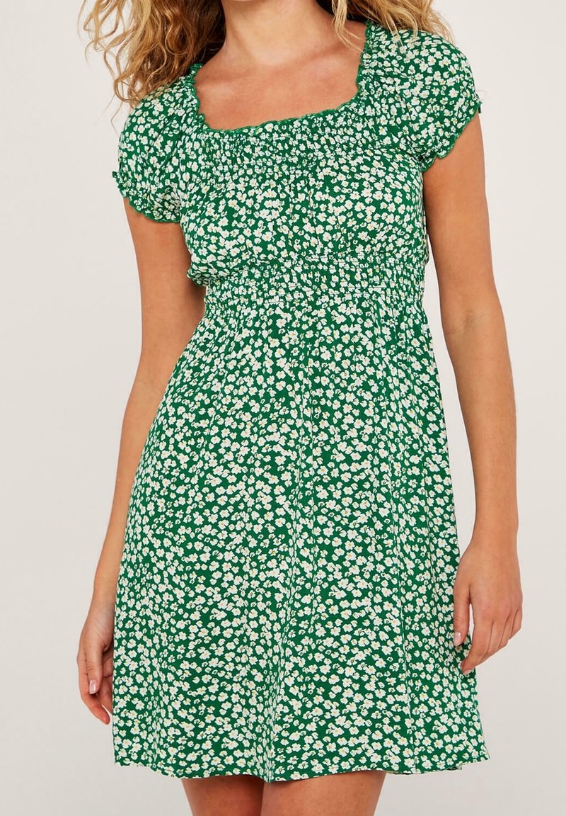 Femme portant une robe verte à manches courtes avec un motif floral blanc et un corsage smocké, debout devant un fond uni.
