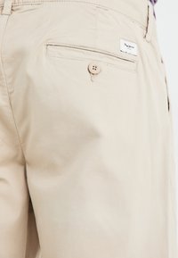 Beige cargo shorts with a buttoned back pocket and a small brand label. The fabric appears smooth with minimal visible texture.