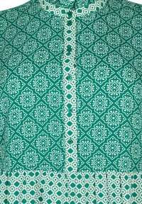 Un tessuto verde a motivi presenta intricati disegni geometrici in bianco. Stilizzato con una chiusura a bottone e colletto a volant, che mette in risalto trame contrastanti.