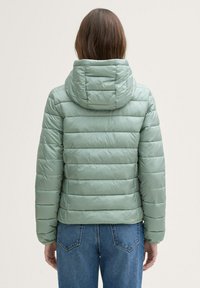 Verstevigde groene jas met een capuchon, met horizontale gestikte secties en een gladde, glanzende textuur. Gepaard met high-waisted jeans.