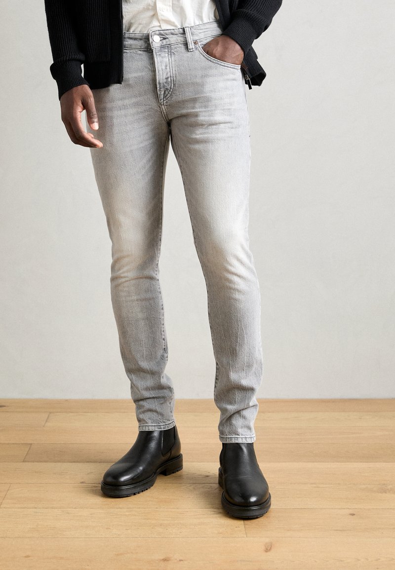 Jeans grigi slim-fit in denim, con una leggera texture sbiadita, abbinati a stivali neri alla caviglia con una finitura liscia e una suola resistente.