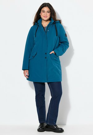 Teal waterproof parka with a hood, button closures, side pockets, and black drawstrings, paired with dark blue jeans and black shoes.