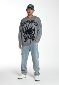 Grauer Pullover mit schwarzem Schädel- und Flammendesign, kombiniert mit hellblauen Jeans und weißen Sneakers. Modell trägt eine blaue Kappe mit Mustern.