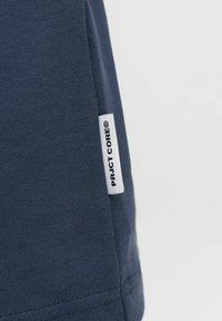 Κοντινό πλάνο σε ύφασμα σκούρου μπλε χρώματος με μια λευκή ραμμένη ετικέτα στη πλευρά, που φέρει μαύρο κείμενο "PRJCT CORE©".