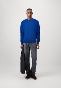 Maglione blu con polsini e orlo a coste, abbinato a jeans grigi e sneakers nere. L'individuo tiene una borsa nera. Design semplice e pulito.