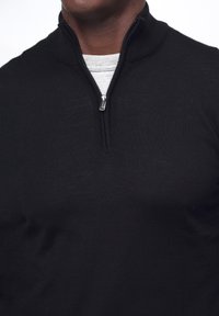 Schwarzer Zip-Kragen-Pullover mit gerippter Textur und hohem Kragen, über einem grauen Hemd getragen, mit einem metallischen Zipper und klaren Linien.