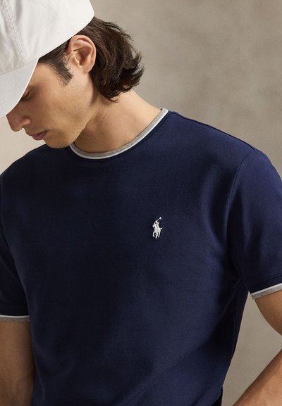 Muž nosiaci bielu čiapku a tmavomodrú košeľu s krátkym rukávom so svetlošedým lemom a malým bielym logom hráča póla na hrudi.