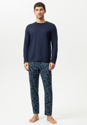 Mann steht barfuß und trägt ein schlichtes dunkelblaues Langarmshirt sowie blaue, mit Blumen bedruckte Hose vor einem einfarbigen weißen Hintergrund.