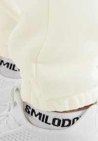 Pantalones de chándal color crema con puños elásticos, combinados con zapatillas blancas y calcetines negros con el logo que muestra "SMILODON". Textura de tela suave visible.
