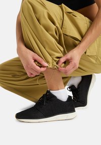 Scarpe da ginnastica nere con tomaia testurizzata e suola bianca, abbinate a pantaloni larghi color khaki. Le mani regolano il cordino dell'orlo dei pantaloni sopra la caviglia.