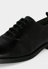 Chaussure Oxford en cuir noir à lacets avec un petit talon et un bout arrondi lisse sur un fond uni.