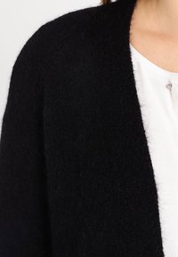 Černý pletený cardigan s jemnou texturou a otevřeným designem, nošený přes bílý top s malým knoflíkovým detailem.
