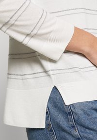 Vit randig tröja med silvertrådar, ribbade mudd och slitsar i sidorna. Matchad med blå denimjeans. Mjuk stickad textur.