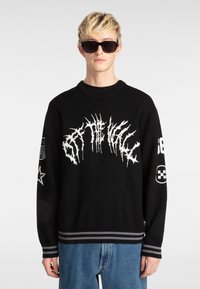 Suéter de punto negro con estampado gráfico blanco, cuello redondo, puños a rayas y dobladillo. Incluye parches de logo en las mangas.