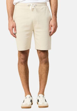 Beżowe szorty z troczkami noszone przez stojącą osobę w białych trampkach z czarnymi detalami na jednolitym tle.