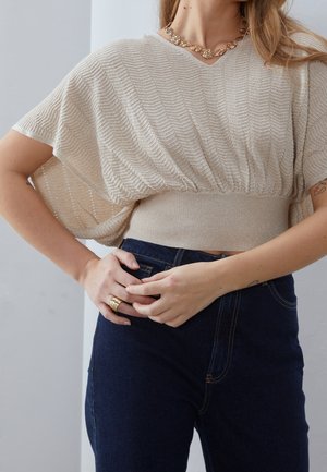 Vrouw draagt een beige gestructureerd kort mouwen top met brede geribbelde tailleband, donkerblauwe high-waisted jeans, gouden ketting en ringen, handen gevouwen.