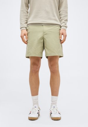 Άνδρας ντυμένος με μπεζ σορτς, μπεζ μακρυμάνικο κορυφή, λευκές κάλτσες μέχρι τον αστράγαλο και λευκά αθλητικά παπούτσια με κόκκινες και μπλε λεπτομέρειες, στέκεται μπροστά σε απλό φόντο.