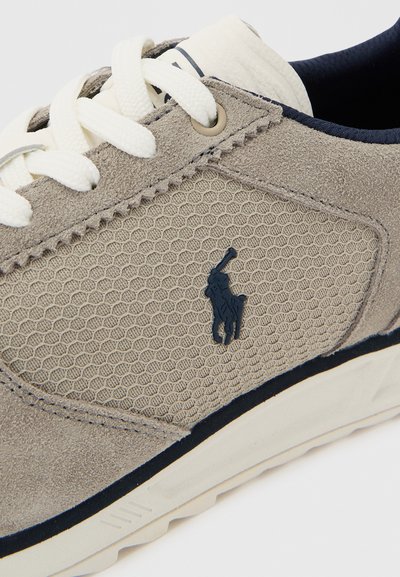 Μπεζ και γκρι αθλητικό παπούτσι με υφή υφάσματος, λευκά κορδόνια και λογότυπο παίκτη πόλο σε σκούρο μπλε στο πλευρικό πάνελ.