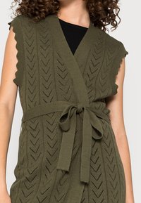 Olivgrön stickad väst med v-ringning, vågiga ärmkanter, knytmidja och chevronmönster. Har ett lätt och texturerat tyg.