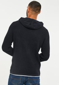 Maglione con cappuccio di un blu scuro e texture, con maniche lunghe, caratterizzato da una vestibilità rilassata e bordi a coste. La vista posteriore non presenta elementi di design.