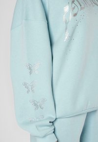 Sudadera de color azul claro con un ajuste oversize, que presenta mariposas adornadas con strass en las mangas y texto decorativo en el frente.