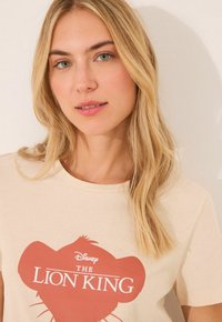 Světle béžové tričko s červenou grafikou siluety lva a textem "Disney KRÁL LVA." Klasický kulatý výstřih, měkký bavlněný materiál.