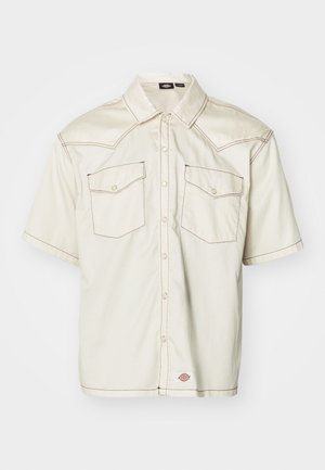 Short-sleeve button-up shirt in beige fabric, featuring two flap pockets, contrasting brown stitching, and a rounded hem.