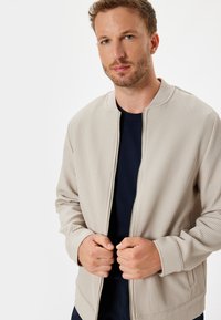 Lys beige ribbet bomberjakke med lynlås foran og rund hals. Har sidelommer og stramme manchetter. Bæres over en marineblå bluse.