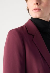 Burgundský blejzr s hladkým texturem, přiléhavým střihem, klopami a strukturovanými rameny, nošený přes černý rolák.
