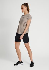 Ljusbeige t-shirt med korta ärmar och en liten logo, kombinerad med svarta åtsittande shorts och svarta sportskor. Slät textur och avslappnad passform.